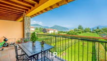 Balcony with a table and chairs, overlooking a lush green garden and scenic mountain landscape. The balcony has a wooden ceiling with exposed beams.