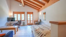 Spacious bedroom with wooden beamed ceiling, large windows providing natural light, tile flooring, leather armchair, desk, and a patterned bedspread on the bed.