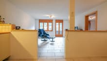 Spacious living room with large windows, wooden furniture, and a balcony or terrace visible through the glass doors.