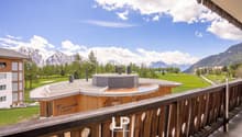 Balcony overlooking a scenic mountain landscape with snow-capped peaks, green meadows, and a fountain or water feature in the foreground. The balcony has a wooden railing and the apartment building appears to be in a resort or alpine setting.