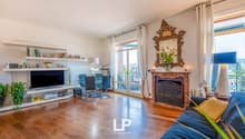 Spacious living room with hardwood floors, large windows, ornate gold-framed mirror, fireplace, TV entertainment unit, and dining area with glass table and chairs