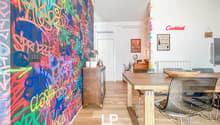 Colorful graffiti-style mural wall, wooden dining table with chairs, shelving unit, and a fireplace visible in the room.