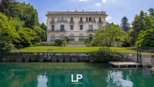 A large, multi-story historic building with a classical architectural style, surrounded by lush greenery and a body of water. The building has balconies, arched windows, and a decorative roofline.