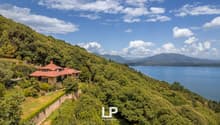 A large, two-story house with a red roof situated on a hillside overlooking a lake and mountains. The house is surrounded by lush, green vegetation and trees. The landscape features a scenic view of the lake and mountains in the distance.