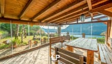 Enclosed terrace, wooden benches and table, glass walls, hanging lamp, view of mountains and water, decorative potted plants on ground