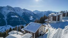 A group of snow-covered houses on a hillside with a ski track, surrounded by mountains and trees. The sun is shining brightly in the sky.
