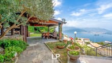 Garden with covered terrace, lake view, stairs to balcony, wooden patio table, chairs, greenery, flowers, olive tree, mountain background