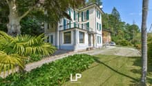 Two-story villa with a balcony, driveway, and parked car. It is surrounded by a well-maintained garden and trees. There are plants on either side of the driveway.