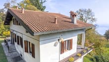 Two-story house with a brown tiled roof, wooden windows, and a tiled patio with plants. The house is raised on the ground floor and has a wooden balcony with a railing. There is a chimney on the left side of the roof.
