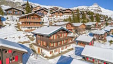 Mountain village, multiple wooden chalets, snow-covered rooftops, surrounded by trees and mountains