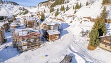 Wooden houses, snow covered mountain, ski lift in the background, trees and shrubs, a house under construction