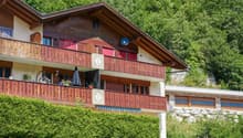 Apartment building, multiple wooden balconies, red shutters, hedge, grassy hillside, stone retaining wall