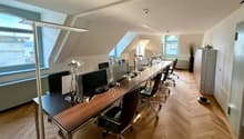 Long desk with multiple computer monitors, office chairs, lamps, wooden floor, white walls, windows, radiators, potted plant