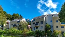 Two multi-story buildings with balconies, yellow exterior, trees, mountains in background, road