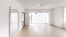 Spacious living room with large windows, hardwood floors, and track lighting. The room has a balcony or terrace visible through the windows.