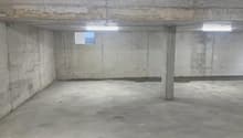 This image shows a large, empty concrete garage or storage space. The walls and floor are made of plain concrete, and there are a few small windows visible. The space appears to be unfinished and utilitarian in nature.