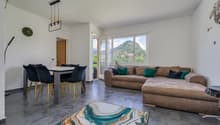 Spacious open-plan living area with large windows offering a scenic mountain view, a comfortable brown sofa, a dining table with black chairs, and a modern light fixture overhead.