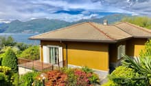 Two-story house with a yellow exterior, a tiled roof, and a balcony overlooking a scenic lake and mountains. The house is surrounded by lush greenery and colorful flowers in the garden.