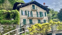 2-story house with a traditional Alpine-style architecture, featuring a sloped roof, wooden balconies, and green shutters. The house is surrounded by lush greenery and situated on a hillside overlooking a scenic landscape.