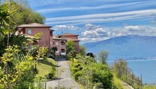 The image shows a group of multi-story residential buildings with a Mediterranean-style architecture, surrounded by lush greenery and overlooking a scenic lake or body of water. The buildings have terracotta-colored roofs and are set against a backdrop of