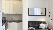 The image shows a small studio apartment with a kitchenette and a living area. The kitchenette has white cabinets, a sink, and a dishwasher. The living area has a black leather sofa, a large wall mirror, and a floor lamp.