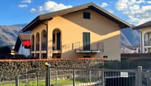 2-story yellow stucco house with arched windows, balconies, and a metal roof. The house is surrounded by a metal fence and landscaping with flowers and shrubs.