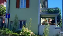 Two-story house with green exterior, red shutters, balcony, and landscaped garden with flowers and plants. The house has a street sign indicating the name 'Beatrice'.