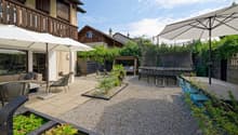 large gravel area, two white umbrellas, a few chairs, a table, plants, trees, trampoline, garden furniture