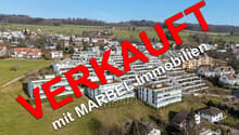 The image shows an aerial view of a town with a mix of residential and commercial buildings. The town is situated in a hilly, forested area with a clear blue sky in the background. The buildings appear to be a mix of modern and traditional architectural s
