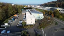 An industrial facility with a large parking area with multiple vehicles parked. A warehouse with a solar panel roof is situated on the property. Several buildings and trees are in the distance.