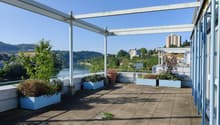 The image shows an outdoor terrace or balcony area with a view of the surrounding landscape. The terrace has a concrete floor and is covered by a metal frame structure. There are several potted plants and flowers placed around the terrace, adding greenery