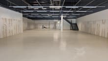 A vacant commercial space with gray concrete flooring, white walls, exposed beams, and overhead fluorescent lighting. There is a staircase in the right corner and an entrance at the back.