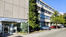 A modern building, likely an office building, with a glass door, name board reading 'Frankenstrasse 70', grey exterior, multiple windows, a few cars parked outside.