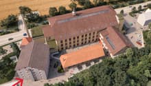 This image shows an aerial view of a large, multi-story brick building with a red tile roof. The building appears to be surrounded by trees and greenery, with a paved courtyard or plaza in front of it. There are several vehicles parked in the area around