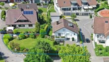 House with green roof, car parked in front, garden, solar panels, large grassy lawn, residential area, other houses and cars visible in the background
