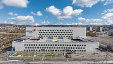 Large modern commercial building with a distinctive white facade featuring a repeating pattern of circular openings. The building has a green roof garden and is situated in an urban setting with other buildings and infrastructure visible in the background