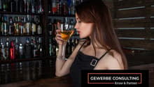 Bar counter with wooden shelf full of bottles, woman sitting on stool drinking a glass of liquor.