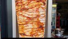 A large piece of meat, possibly chicken, is being cooked on a vertical rotisserie. The meat is sliced into thin layers and is being cooked with a red sauce. The meat is being cooked in a commercial kitchen with a stainless steel oven and a refrigerator in
