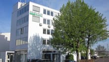 Modern white office building with glass windows, located in Anslagpark, multiple cars parked in front.