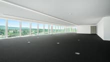 Large open office space with floor-to-ceiling windows offering a panoramic view of the surrounding landscape. The room has a black carpeted floor and recessed lighting in the ceiling.