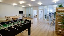 Large open office space with hardwood floors, white walls, and large windows. The space includes a foosball table, a dining table with chairs, and various potted plants. There is also a kitchen area visible through the doorway.