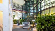 modern lobby, gray couch, white tables, large floor lamps, white wall, gray flooring, large plants, glass windows, staircase