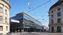 Large glass and steel commercial building with Credit Suisse branding, surrounded by historic ornate buildings, tram lines, and pedestrians on the street