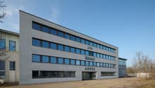 office building, grey, large windows, multiple floors, entrance with text, adjacent parking lot