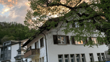 white facade house with brown wooden shutters, balcony on first floor, red roof, mountain in background