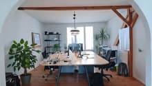 large room with wooden floor, two long tables, office chairs, desk, plant, desk lamp, framed picture, black bag, trolley, ceiling lamp