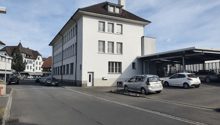 White building, two stories, multiple windows, parking in front, cars parked under carport.
