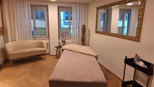 Room contains a chair, a table with a flower vase, a small bed with a fur blanket, and a large mirror