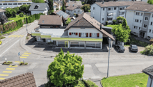 2 story house, brown roof, yellow facade, 2 cars parked in front, street with yellow and white markings, trees and bushes