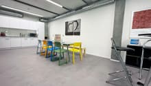 Open room with dining table and chairs. Light gray floors, white walls. Kitchen area with microwave, cabinets, bottles, kettle and a coffee machine. Bar stools next to a table with books and a framed art.